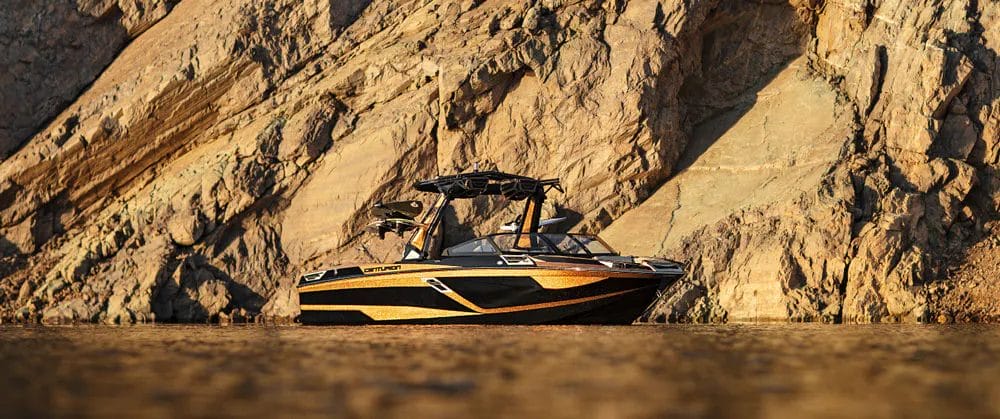 A black and yellow wakeboat in the water near a rocky cliff.
