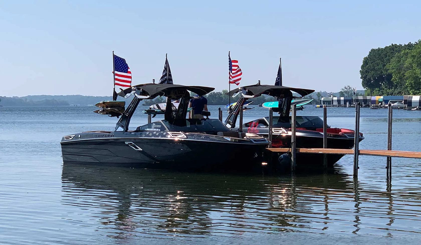 Two wakesurf boats docked at a dock with American flags.