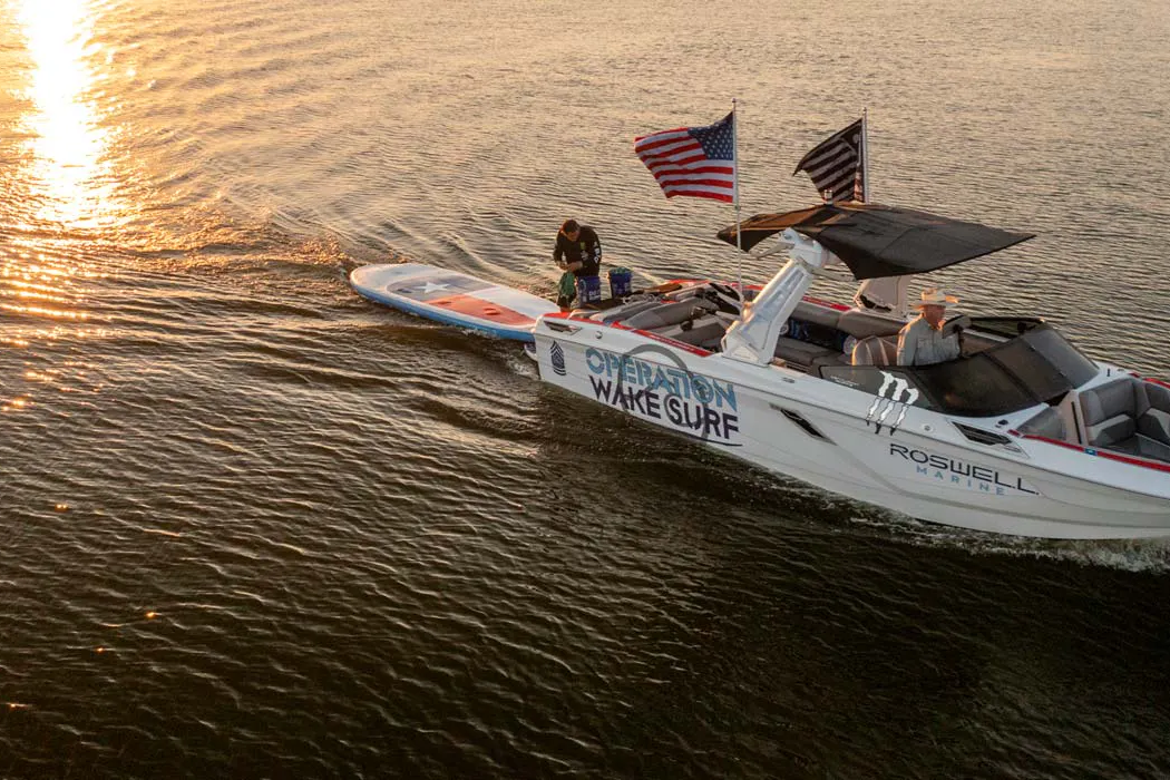 A group riding a wakesurf boat in the water at sunset.