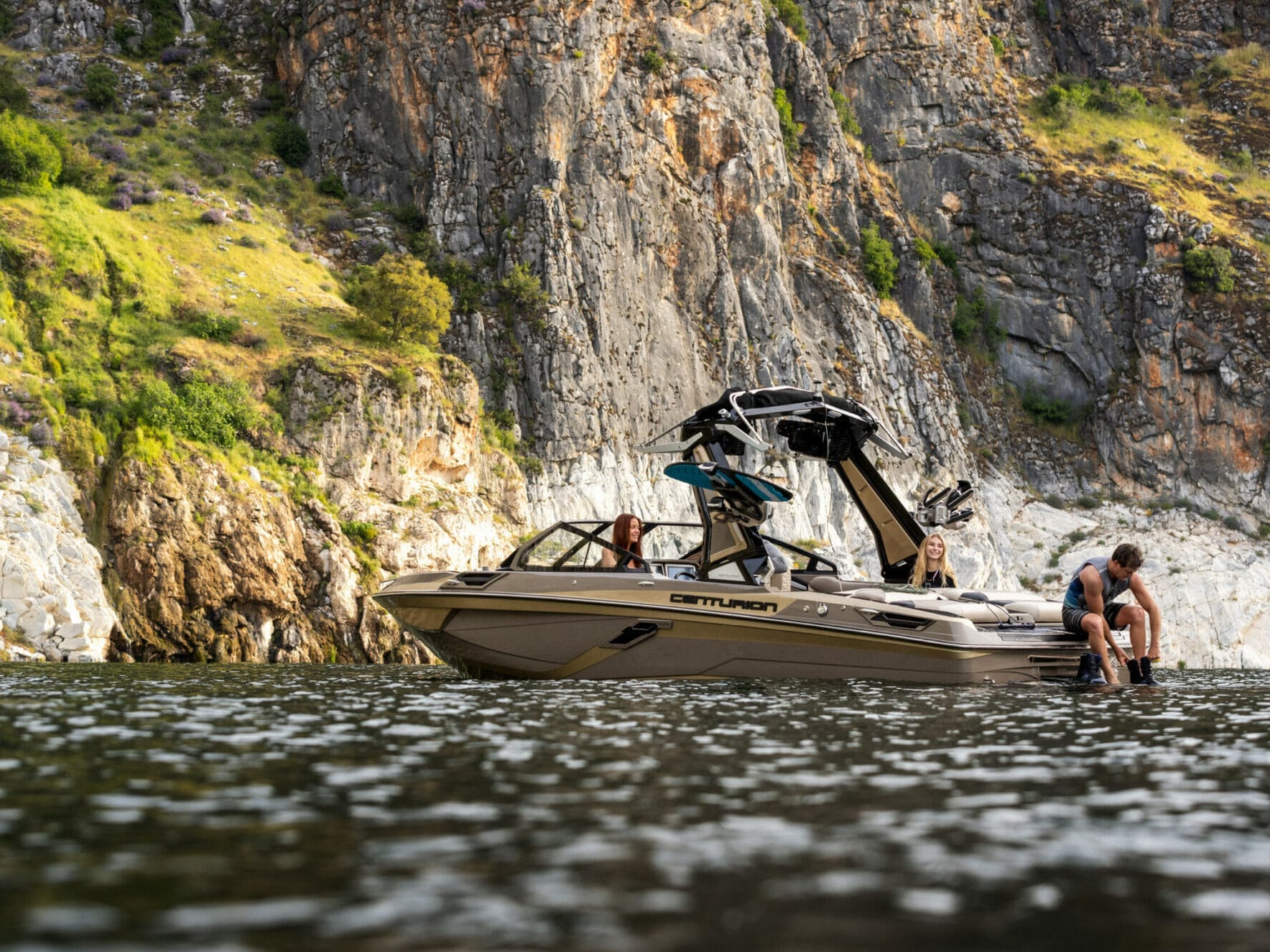 A man is standing on a wakeboat in the water next to a cliff.