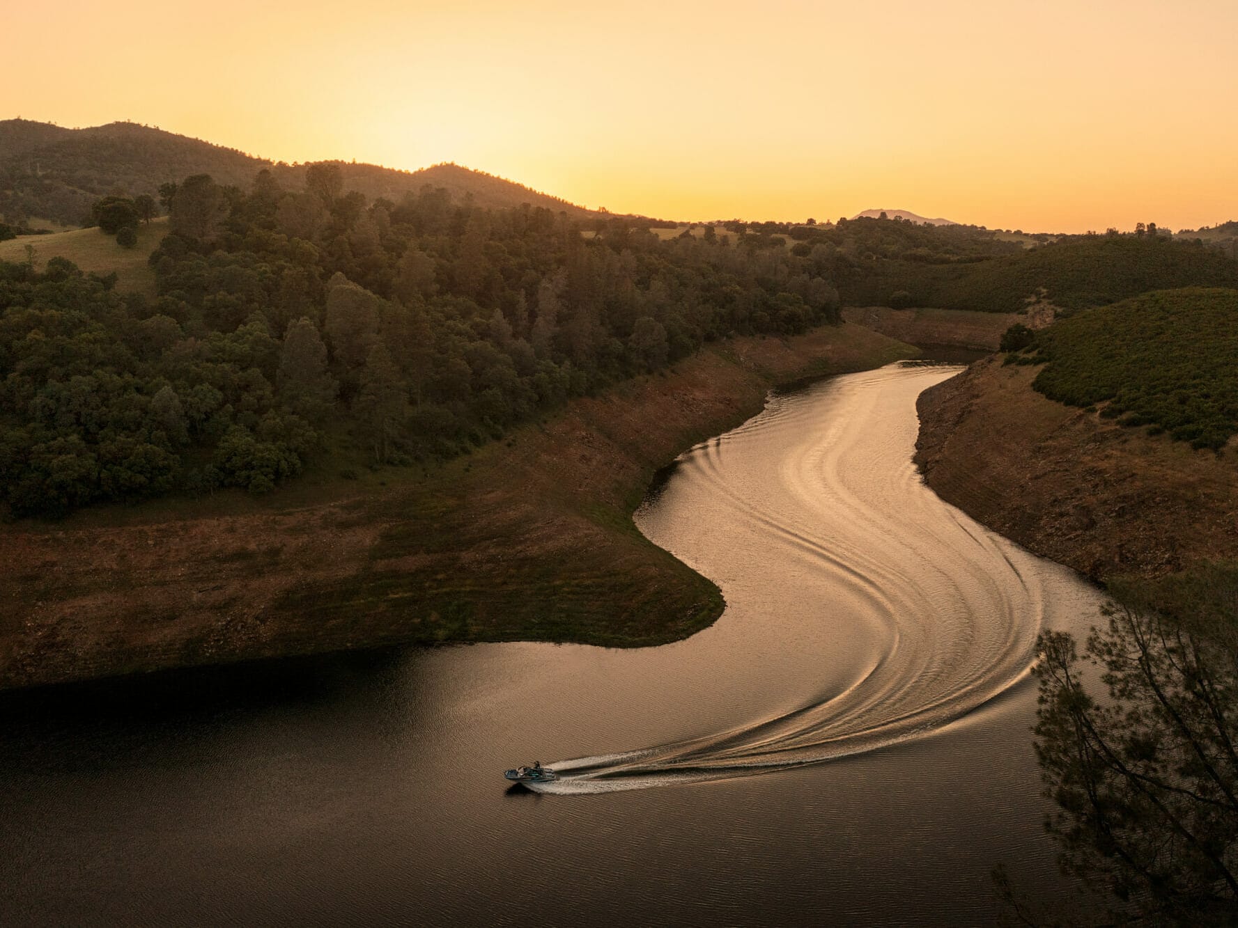 A wakesurf boat cruising down a river at sunset.