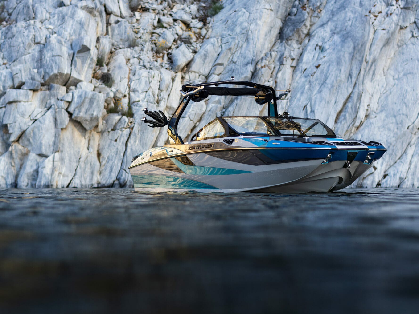 A wakesurf boat is floating in the water near a cliff.