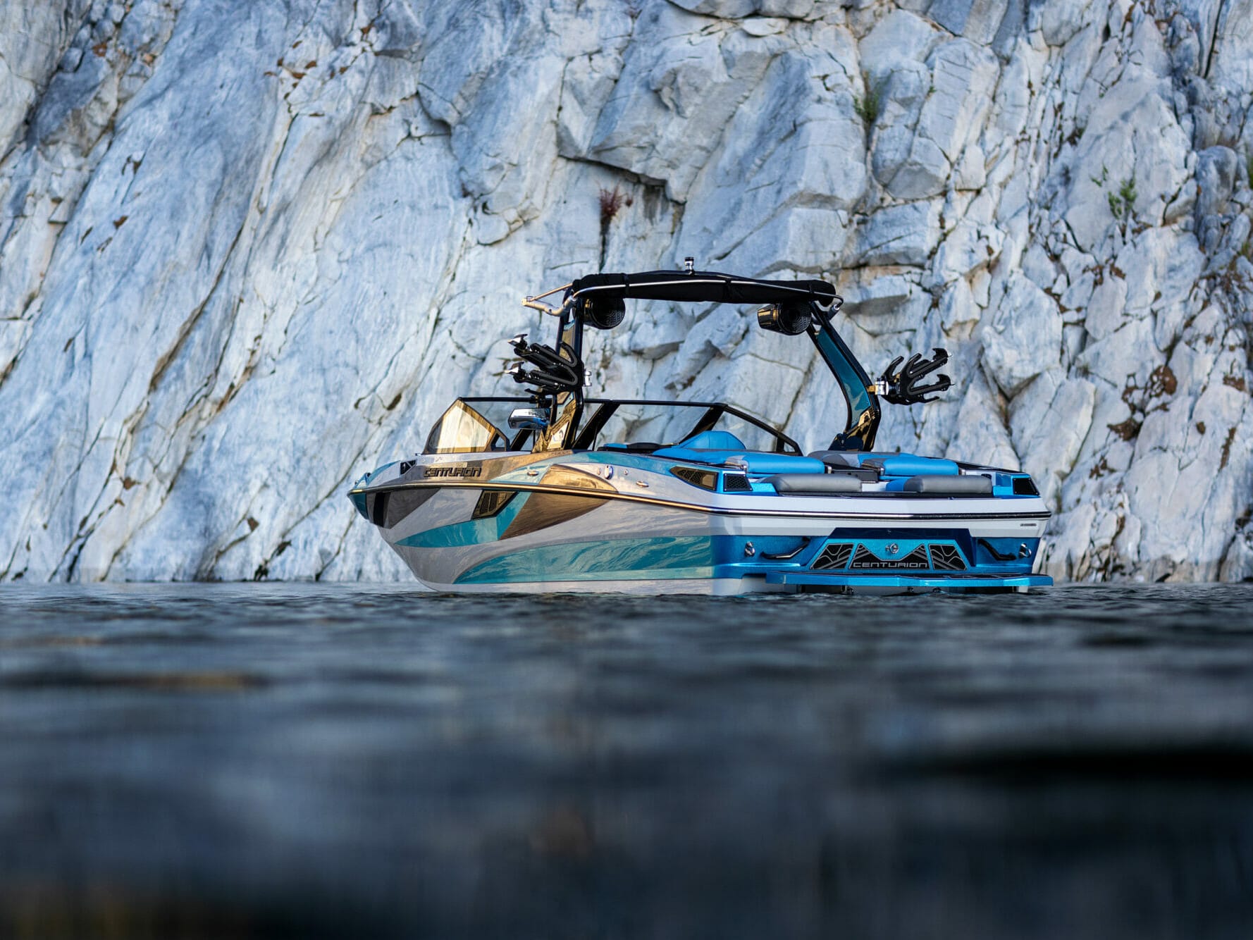 A blue and white wake boat is in the water near a cliff.