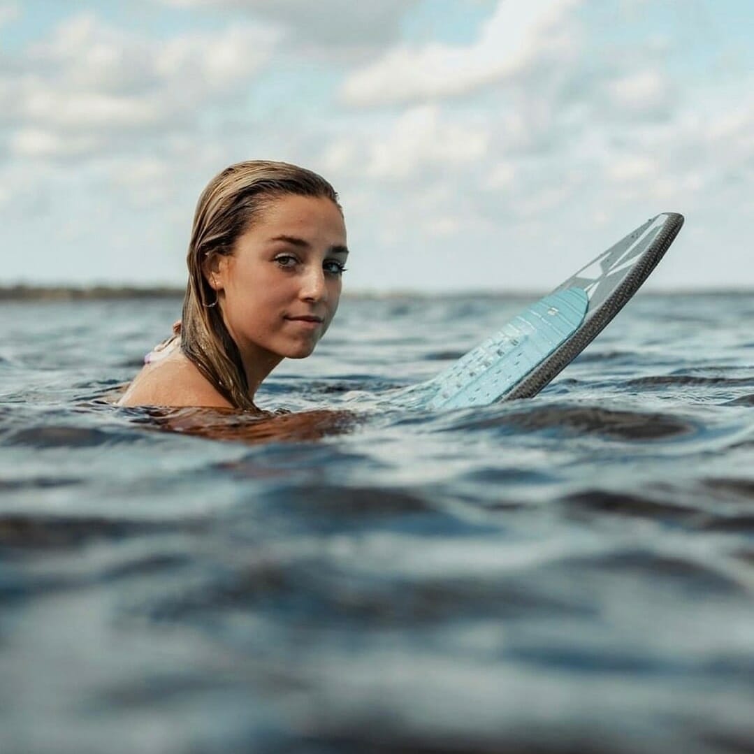 A young woman holding a surfboard in the water near a wakesurf boat.