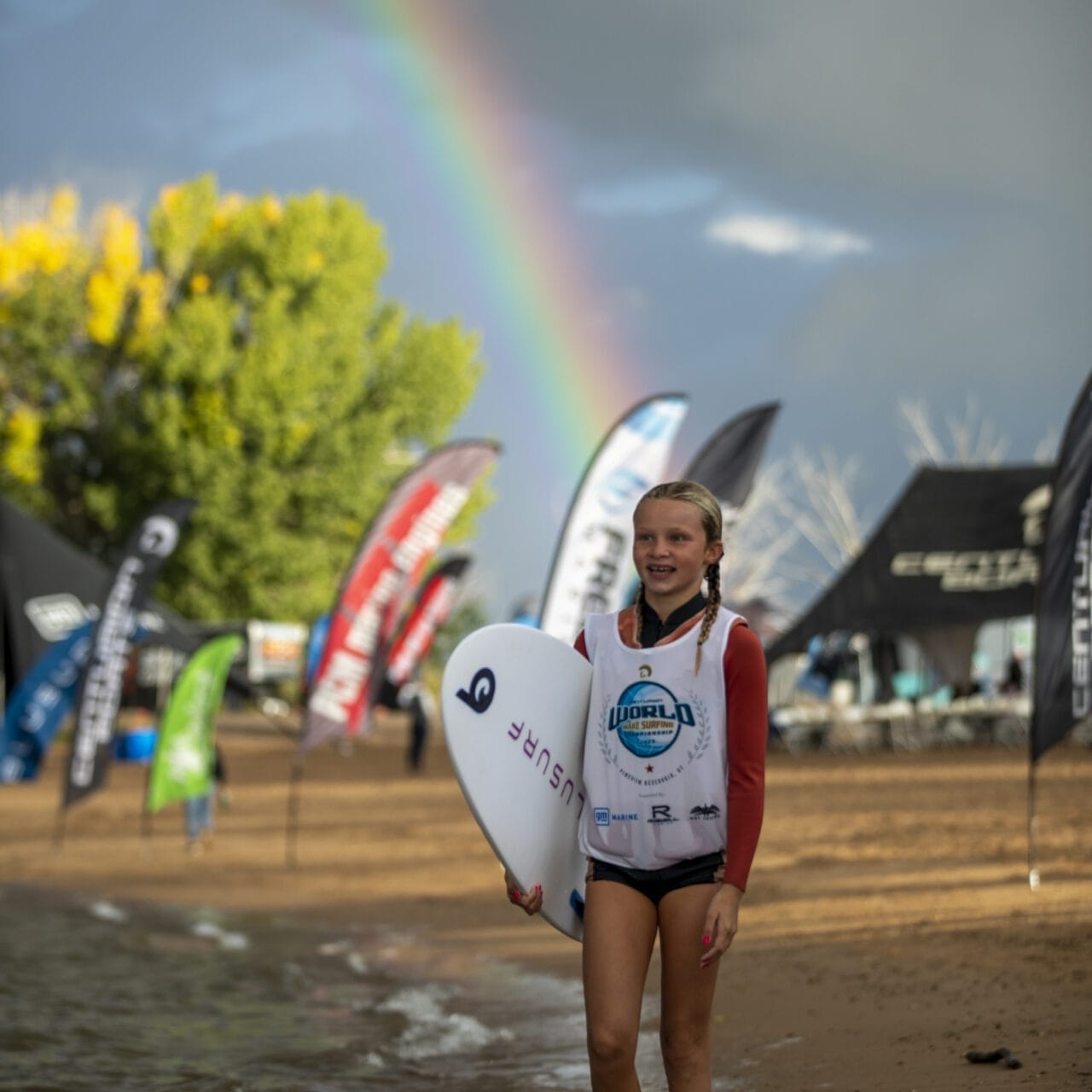Kenzie Hickey, a girl, holding a surfboard in the water with a rainbow in the background.