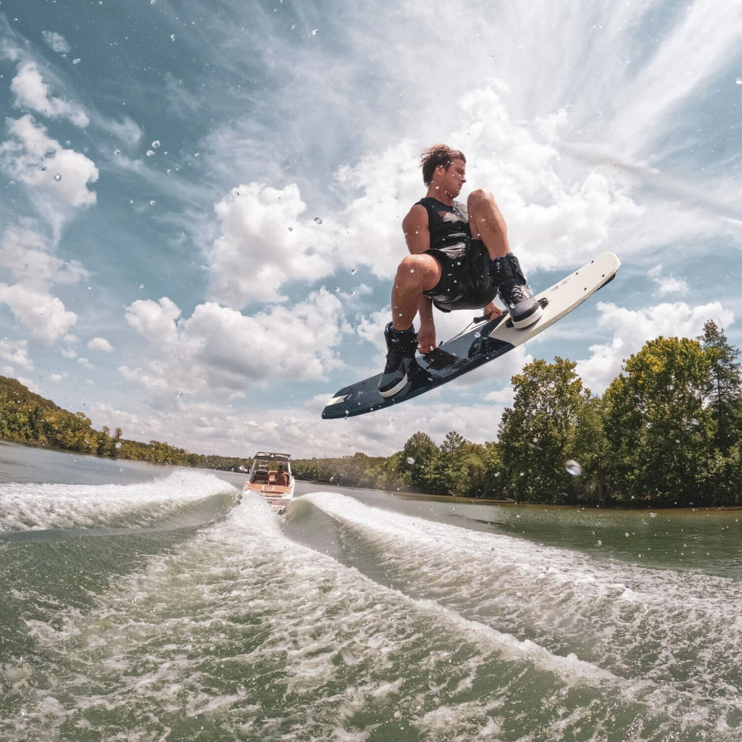 A man is riding a wakeboard behind a wakeboat on a lake.