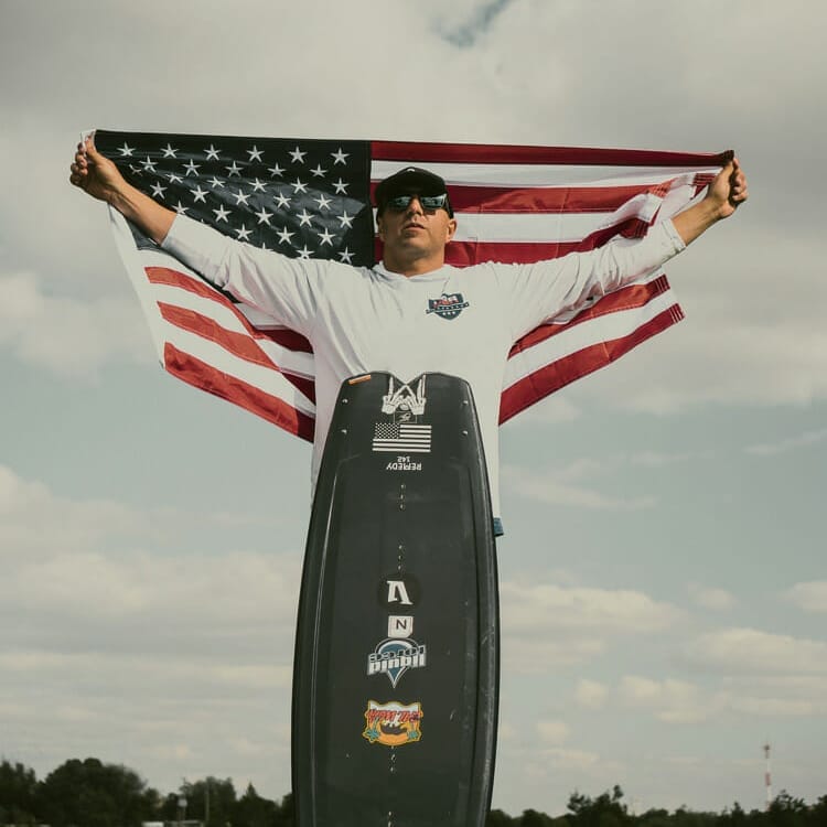 A man holding an American flag and a wakeboard on a wakesurf board.