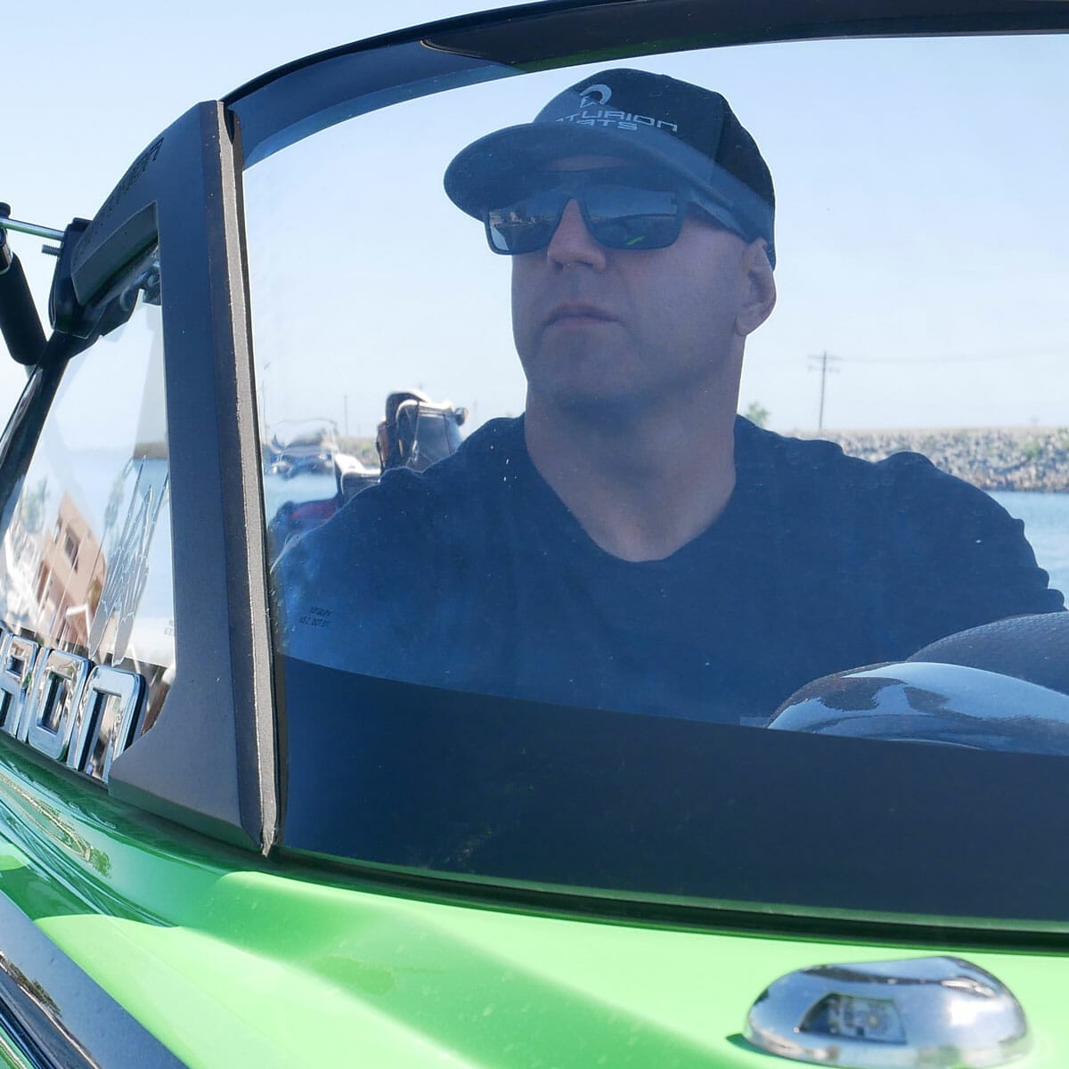 A man in sunglasses is driving a green wakesurf boat.