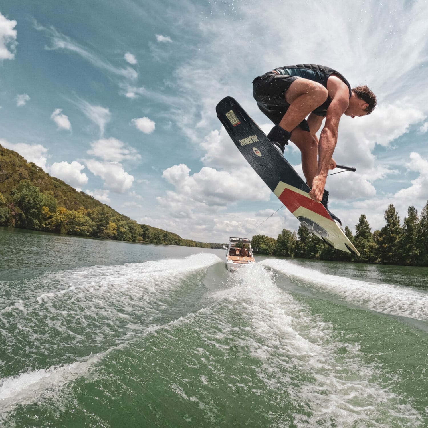 A man is riding a wakeboard behind a wakeboat on a lake.