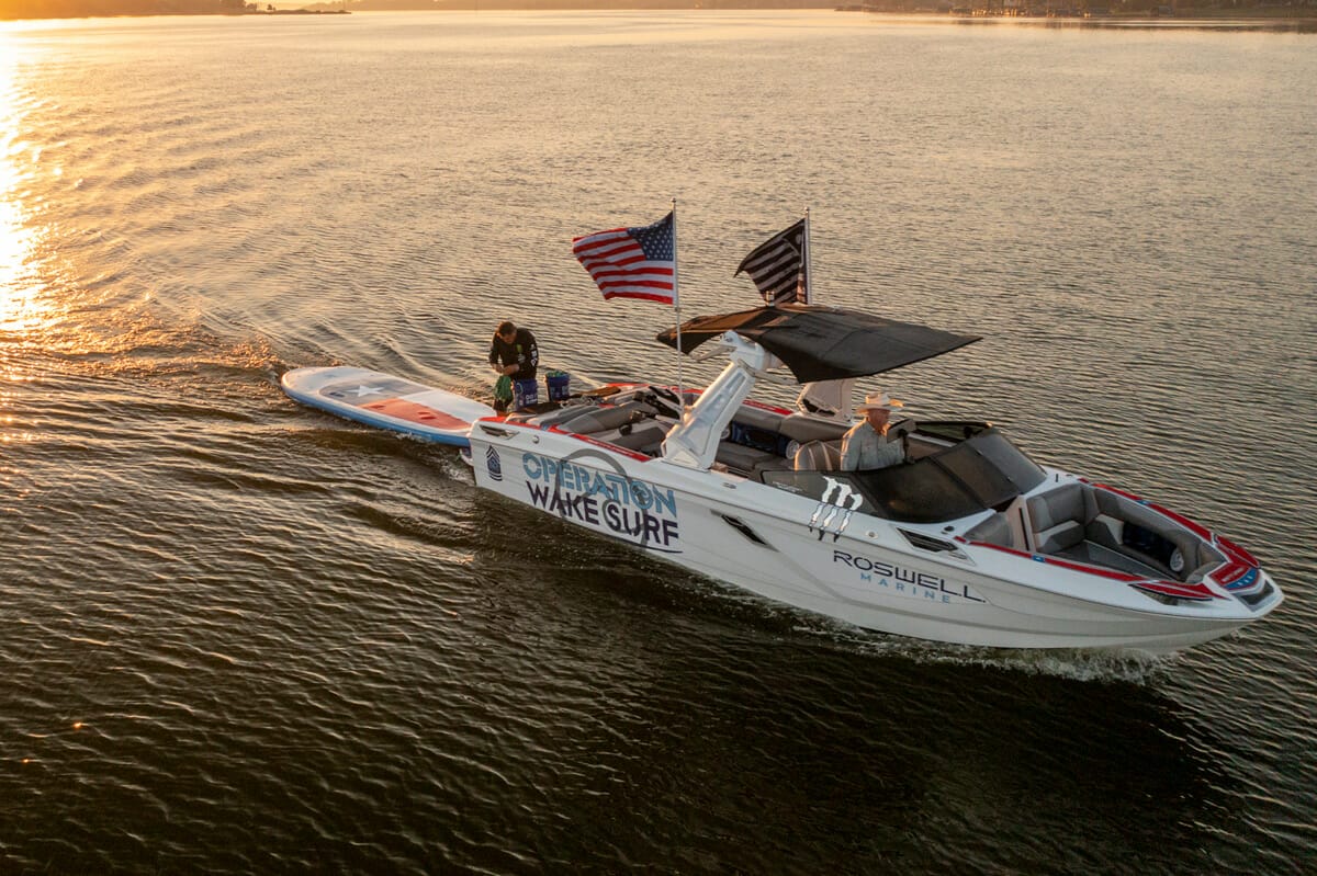 A wakesurf boat with two people on it at sunset.