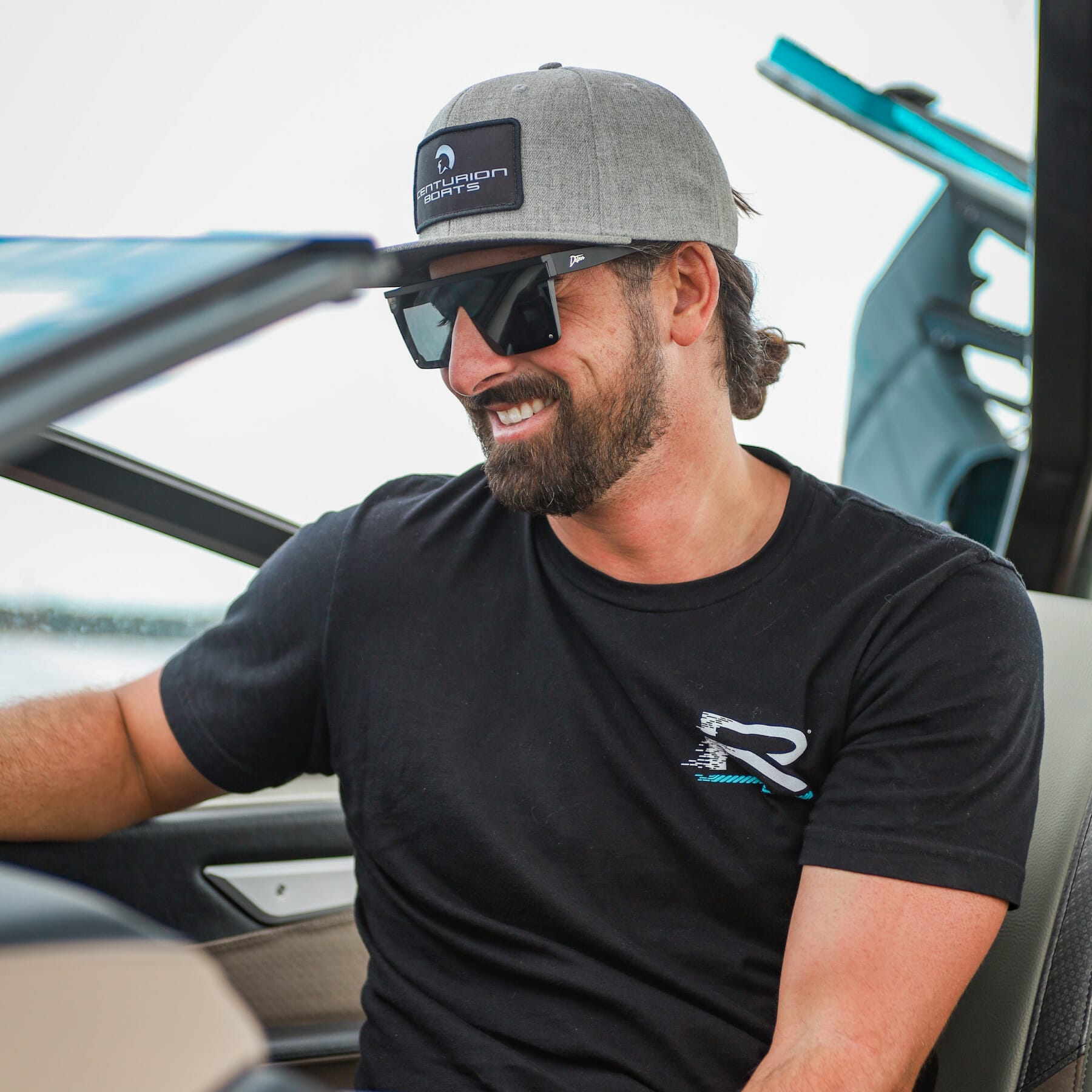 A man wearing sunglasses and a hat sitting in the driver's seat of a wakesurf boat.