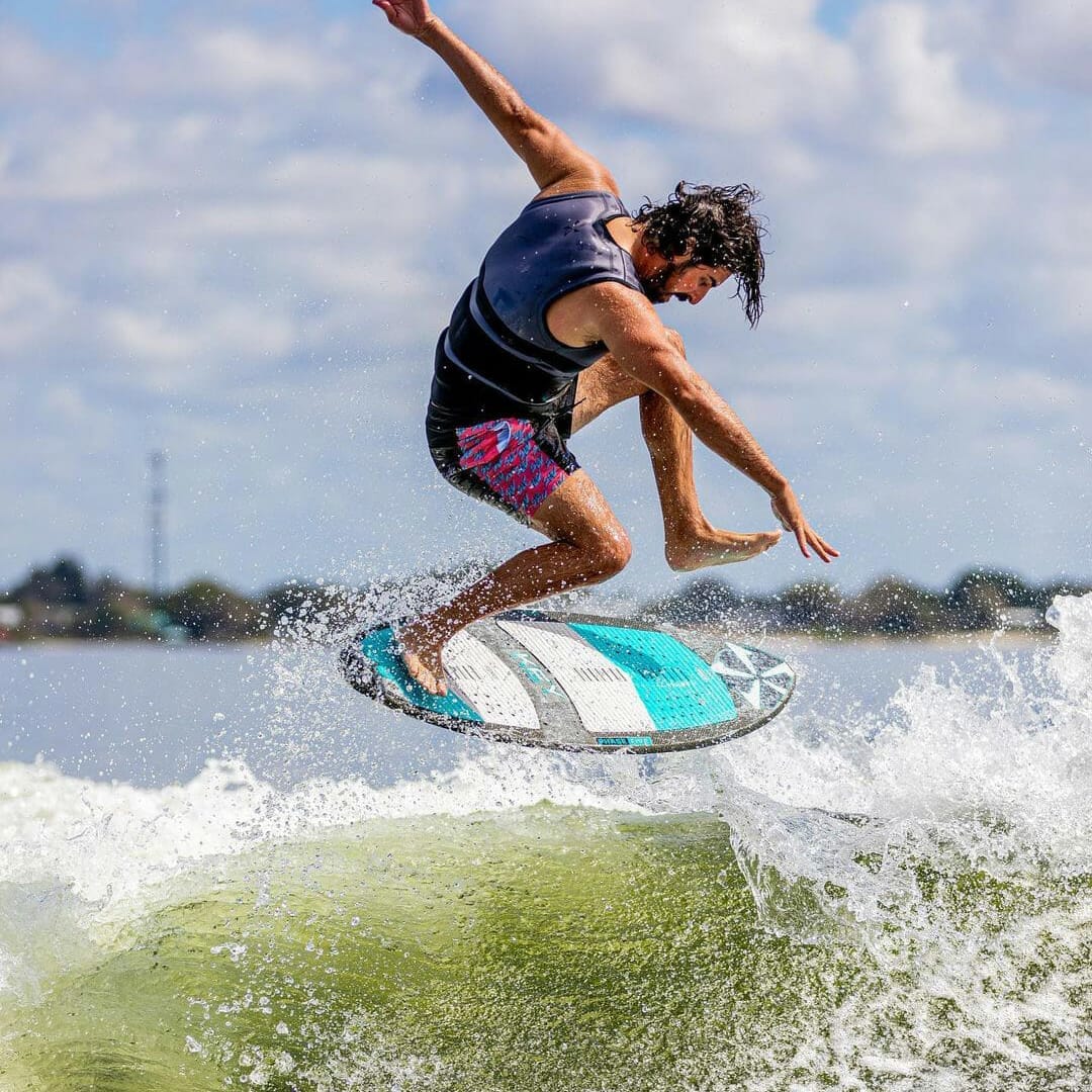 A man is riding a wave on a surfboard behind a wakesurf boat.