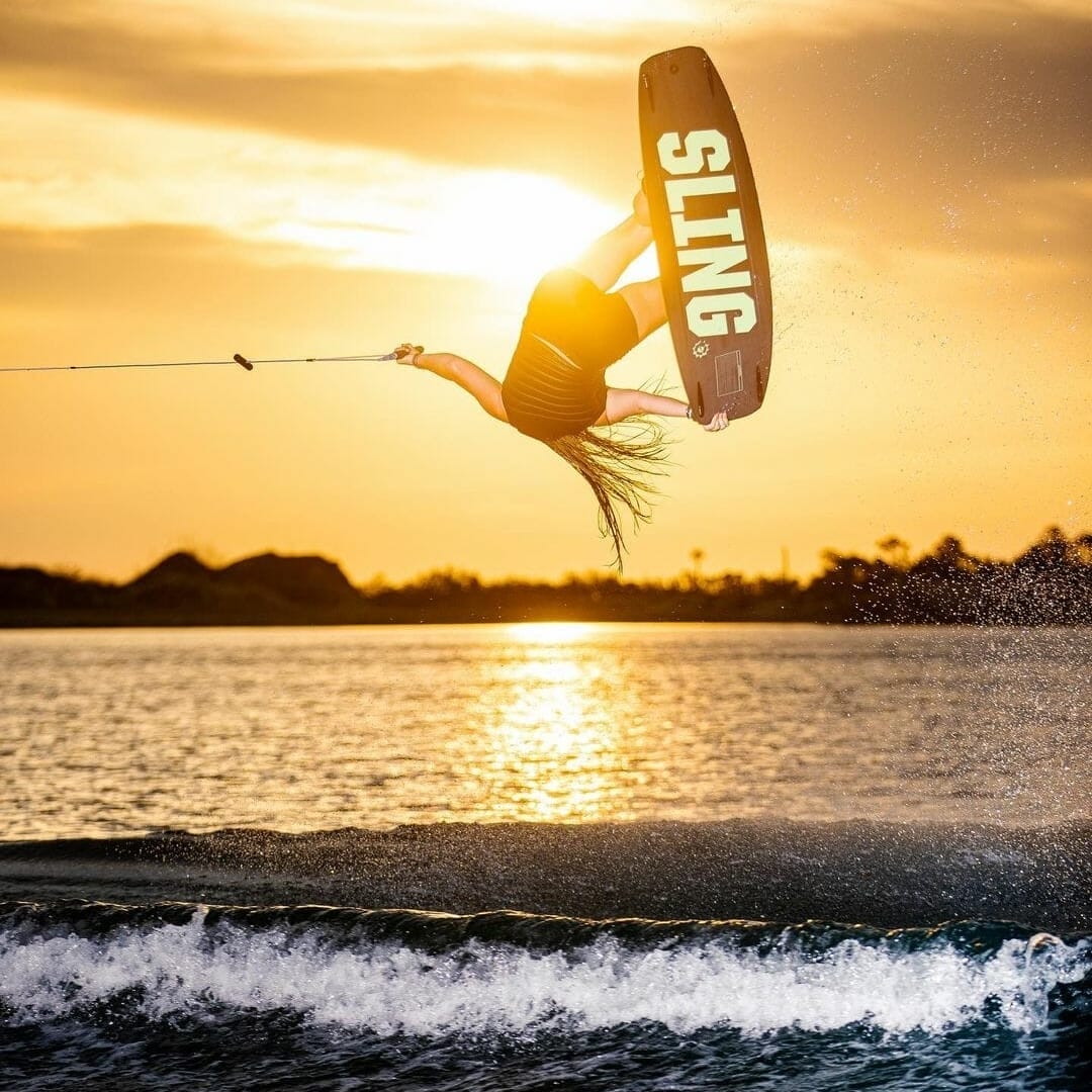 A woman wakesurfing behind a boat at sunset.