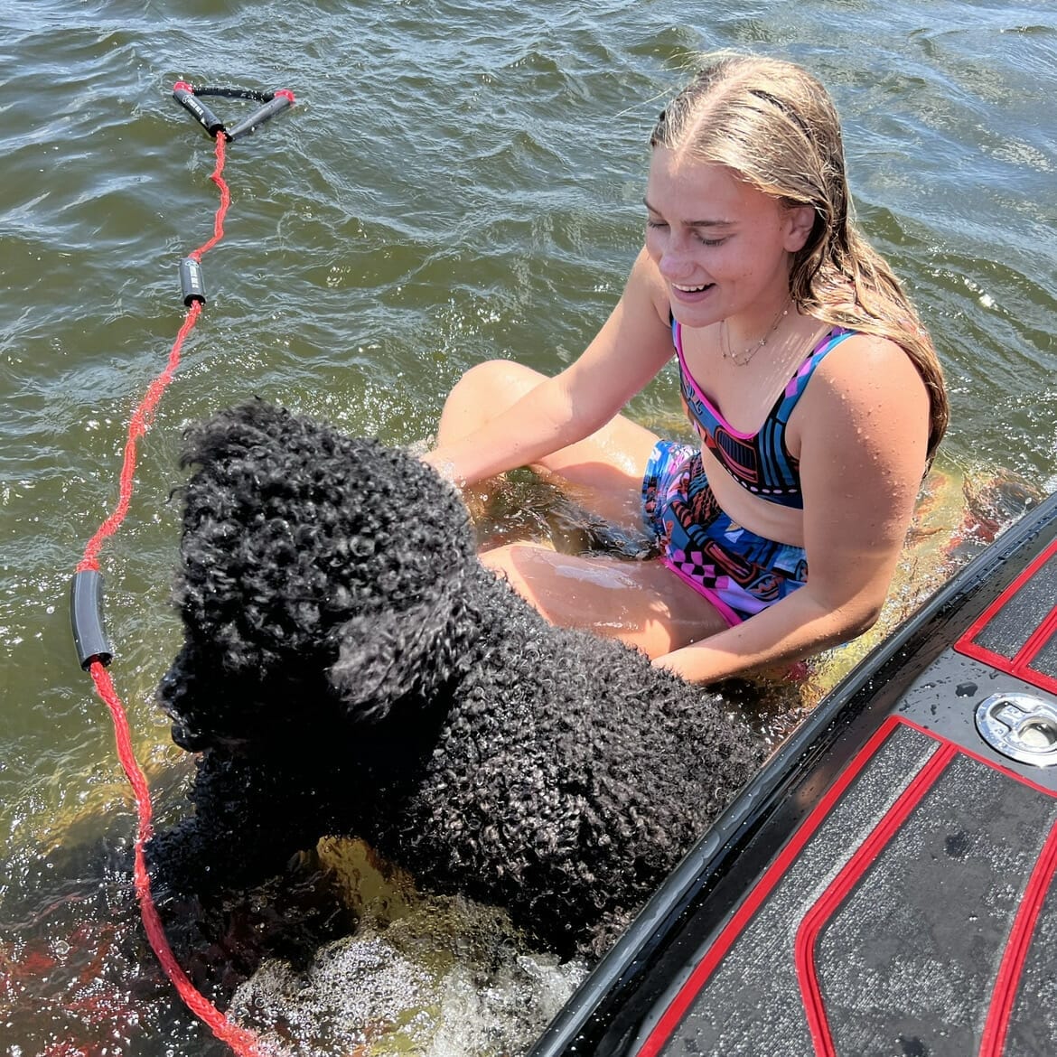 A girl with a black poodle enjoying the water while wakeboarding behind a wakesurf boat.