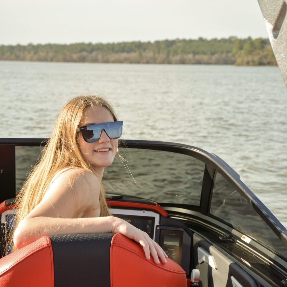 A woman in sunglasses sitting in the back of a wakesurf boat.