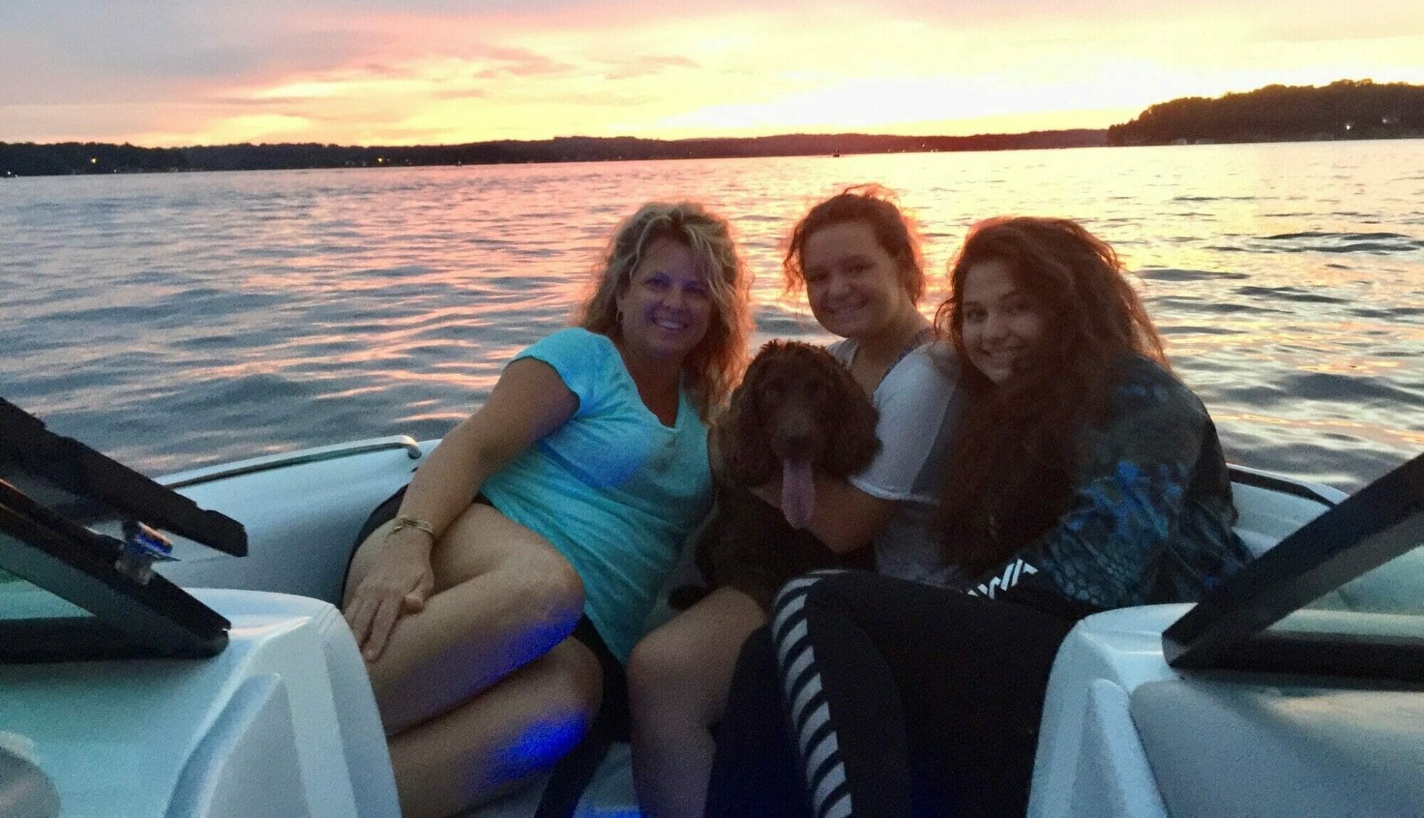 Three women and a dog on a wakesurf boat at sunset.