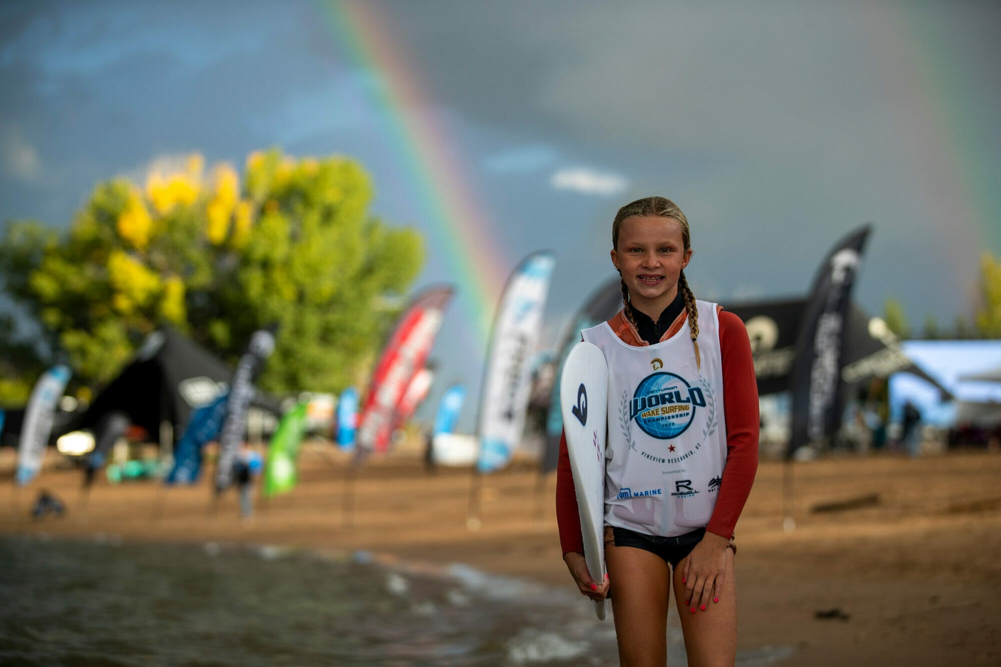 A girl with a wakesurf board standing on the beach with a rainbow in the background.