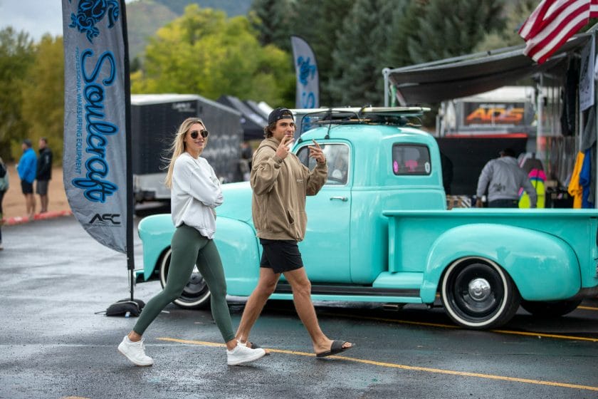 Two women walking in front of a blue truck with wakesurf boards.