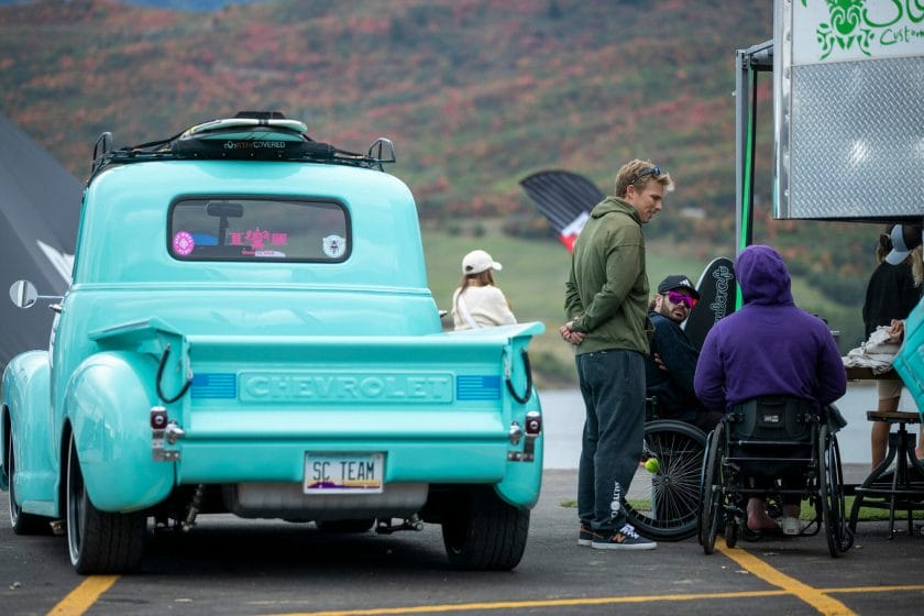 A turquoise truck with a man in a wheelchair next to it, carrying a wakesurf board.