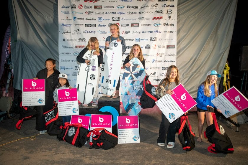 A group of girls standing on a stage during the 2024 WWSC Day 2 Recap, showcasing their surfboards.