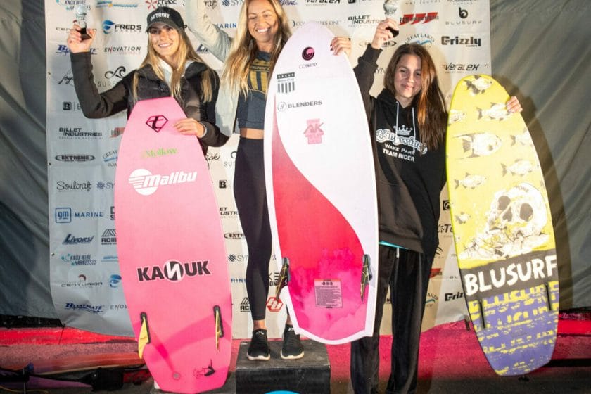 2024 WWSC Day 2 Recap: Three women triumphantly pose on podium with surfboards.