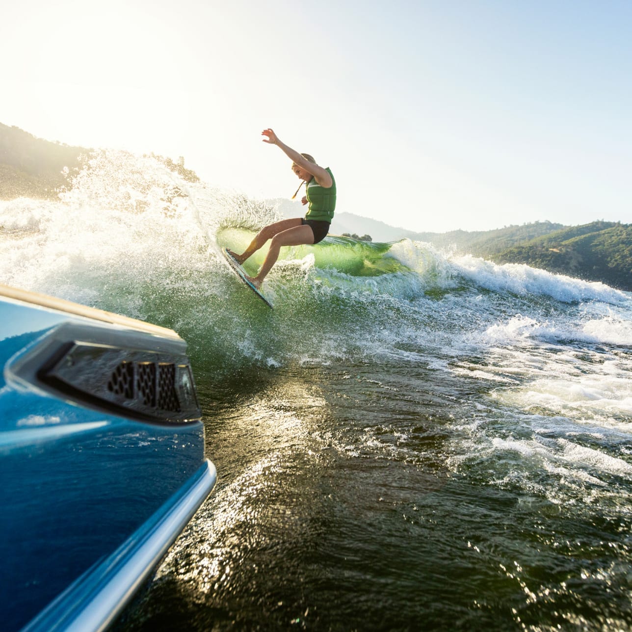 A person wakeboarding behind a Centurion Fe Series boat on a sunny day, with mountains and a clear sky in the background.