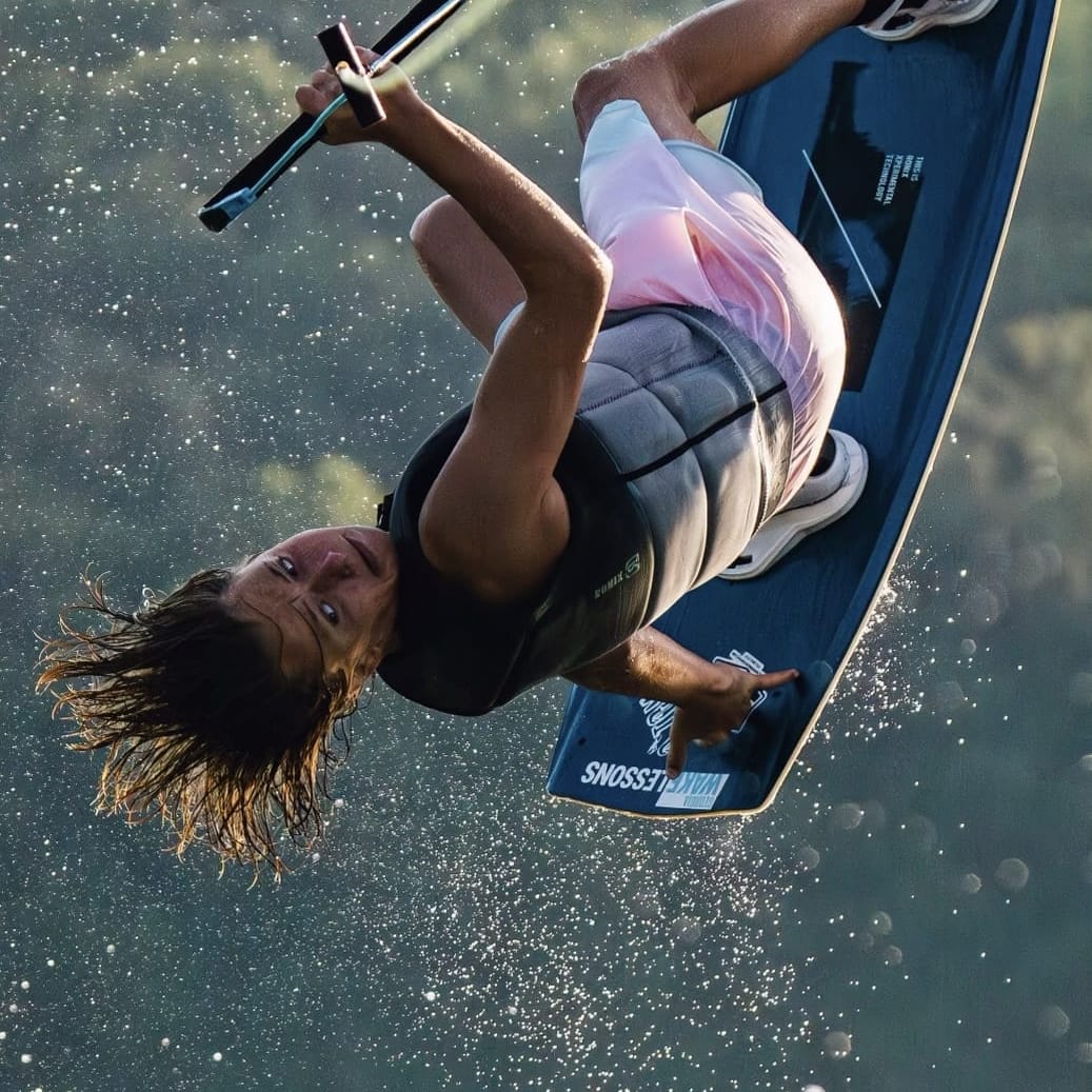 A wakeboarder performs an airborne trick, flipping upside down with a wakeboard attached to his feet, water droplets in the air.