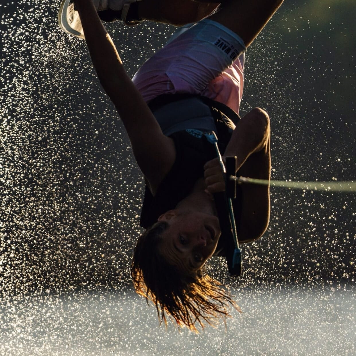 A wakeboarder performs an inverted trick, suspended in mid-air above water with droplets glistening in the sunlight.