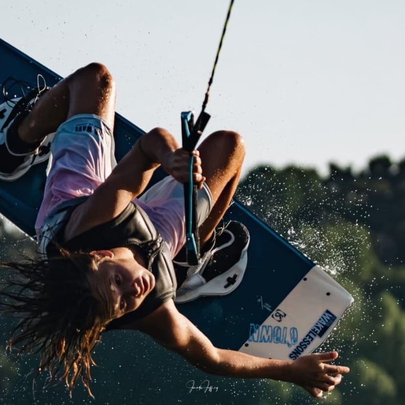 A wakeboarder executes a high-flying trick over a lake, gripping the tow rope with one hand while flipping the board overhead.