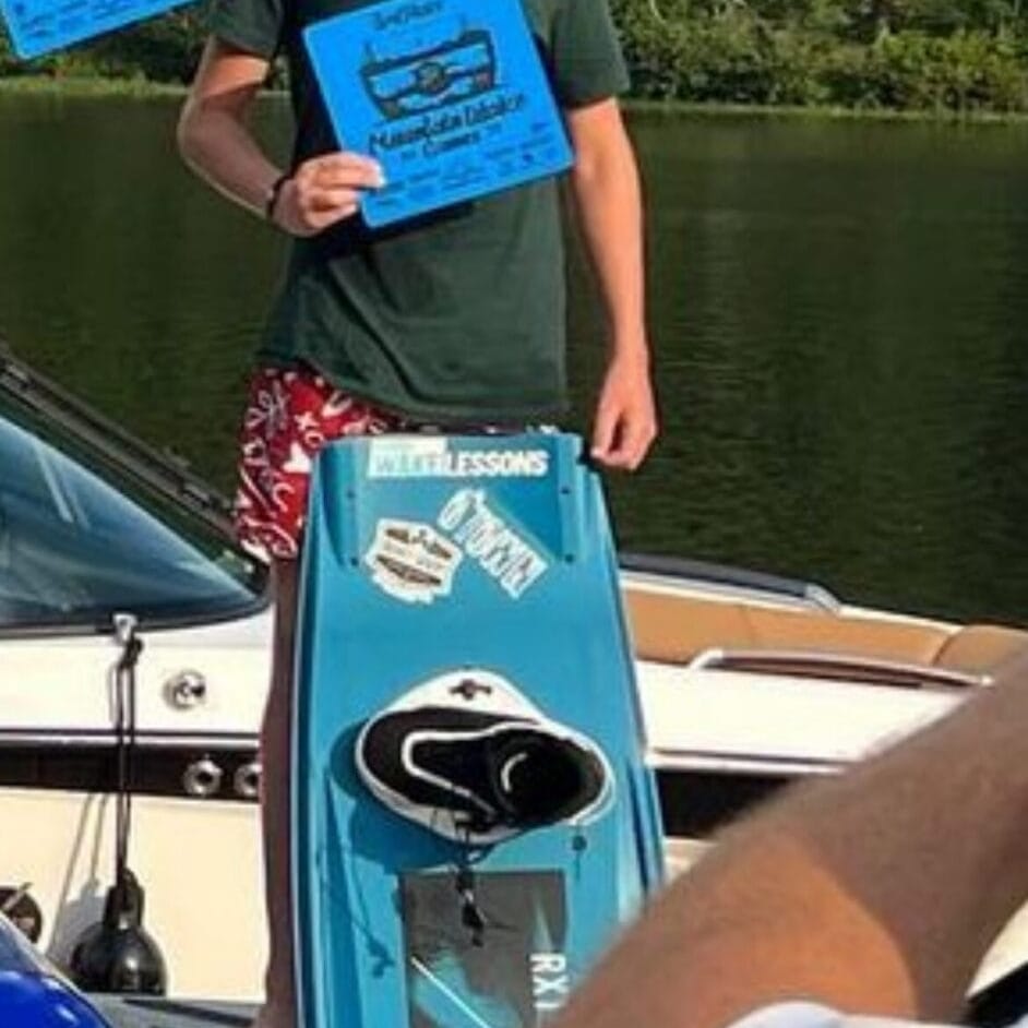 A young man holding two blue certificates stands on a dock with a turquoise wakeboard.