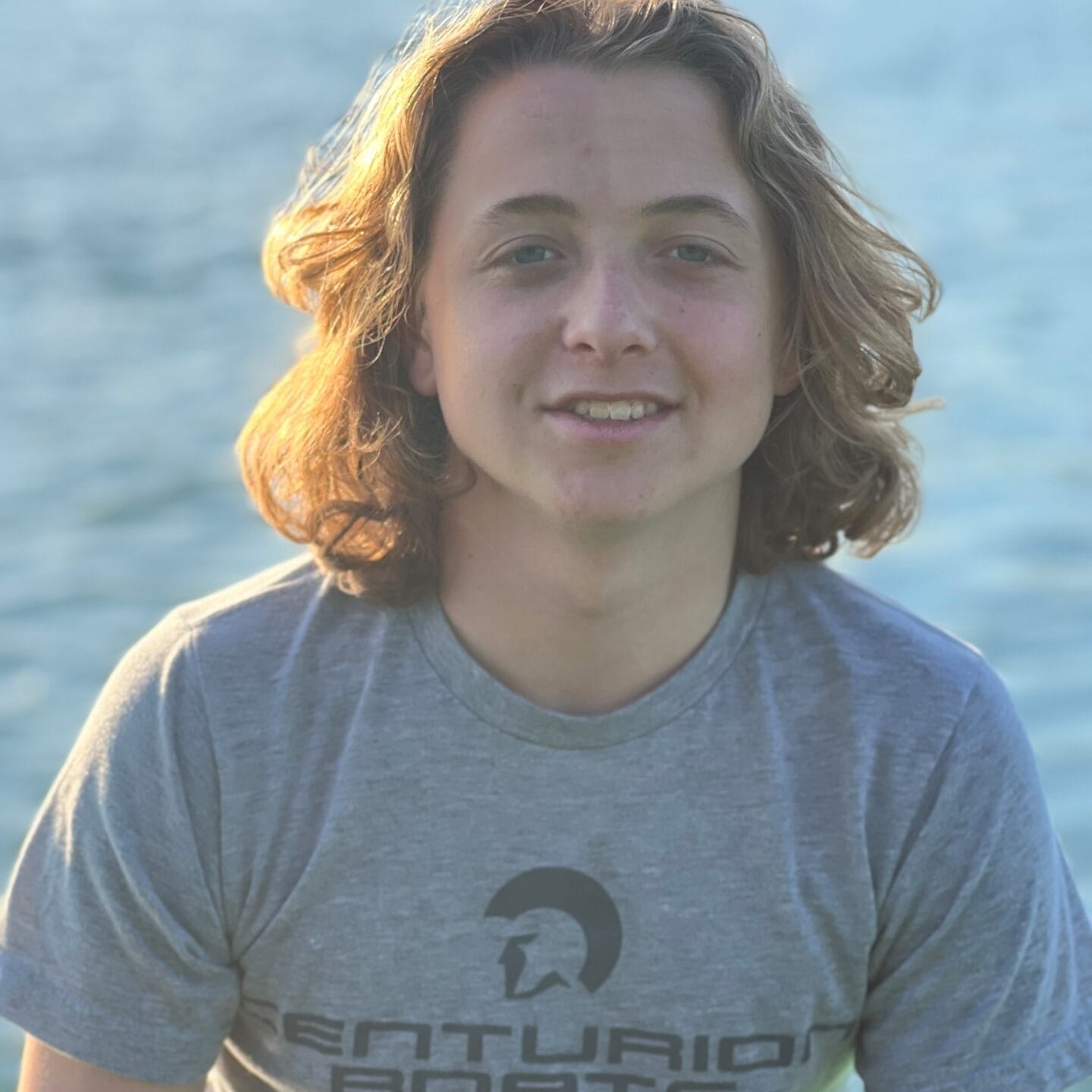 Young man named Camden Marsden with shoulder-length curly hair, smiling, wearing a gray t-shirt, with a lake in the background during sunset.