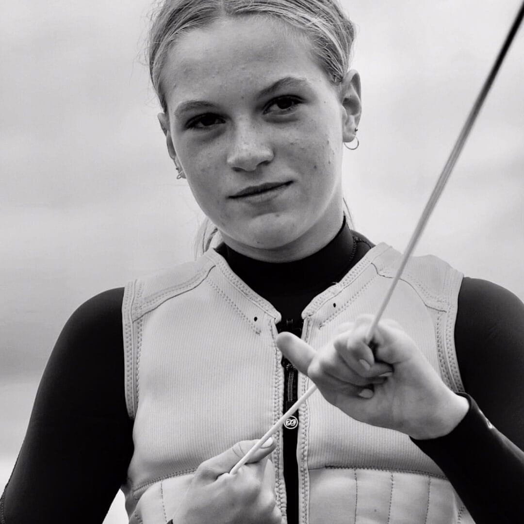 Young woman in a life vest and wetsuit holding a rope, standing by the water, looking directly at the camera.