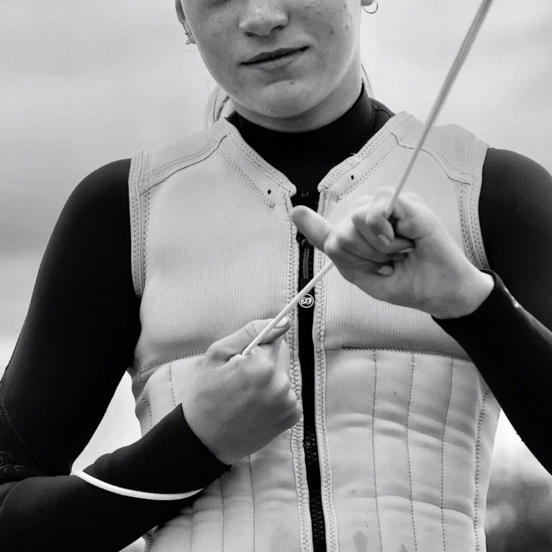 Young woman in a life vest and wetsuit holding a rope, standing by the water, looking directly at the camera.
