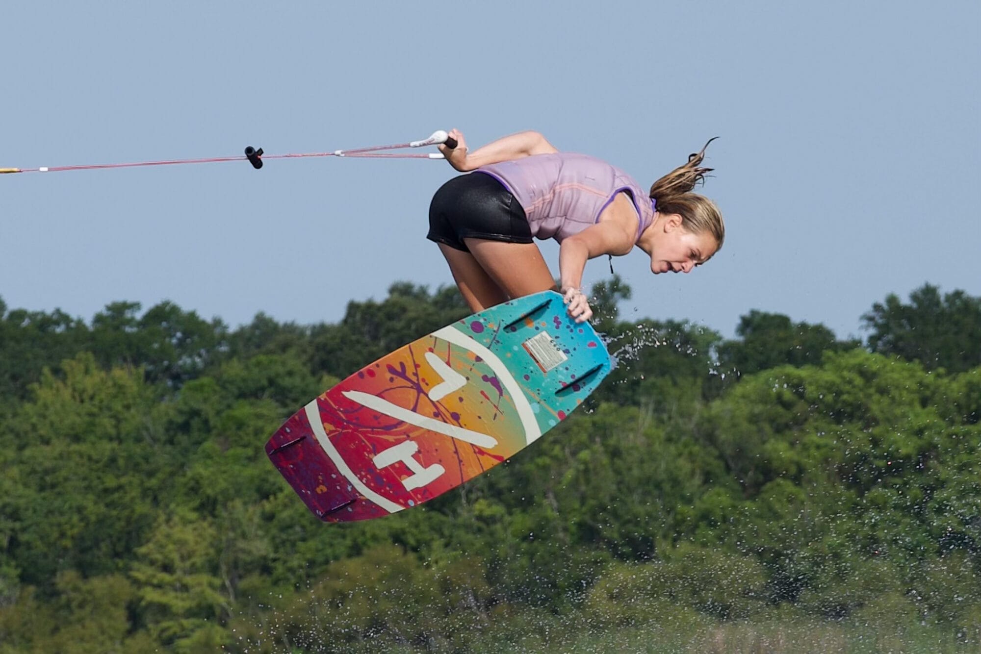 A woman wakeboarding mid-air with a colorful board, holding a tow rope, above a body of water.