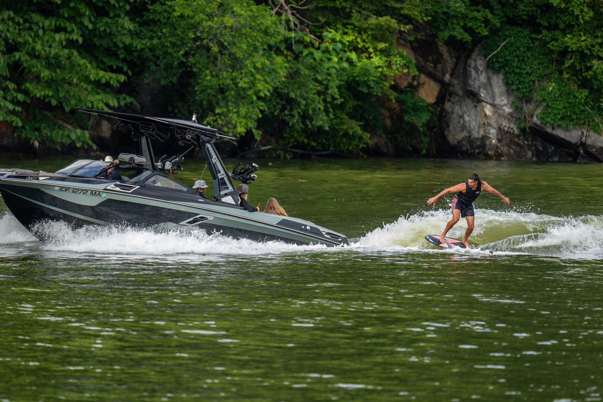 A person is wakesurfing behind a black boat with several passengers on a green, tree-lined body of water.