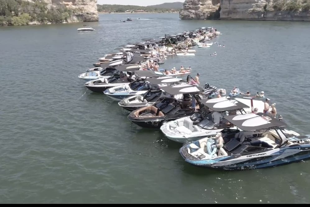 A large group of boats is lined up on a lake, forming a semi-circle. The boats are filled with people enjoying the water and sunny weather, with cliffs and trees visible in the background.