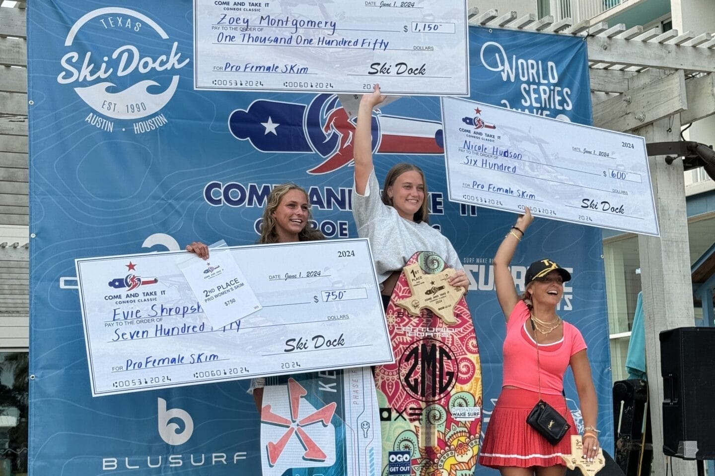 Three female surfers stand on a podium holding large prize checks at a surfing competition. They are smiling, and a surf-themed backdrop displays sponsors' logos.