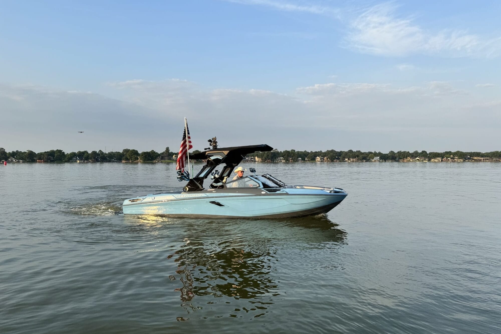 A light blue motorboat with an American flag is moving on a calm lake under a clear sky with some clouds. Trees and houses are visible on the distant shoreline.
