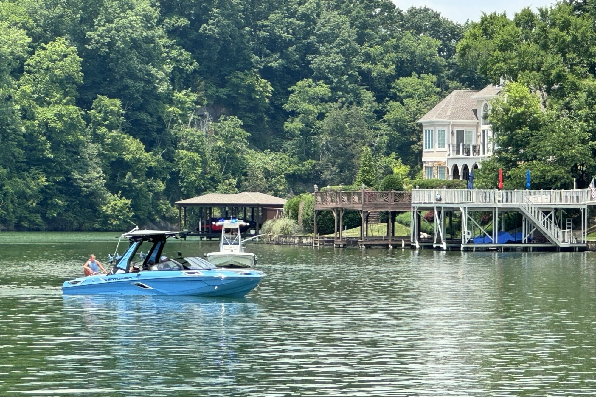 A blue motorboat with several people onboard cruises on a lake near a large lakeside house with a dock and surrounding greenery.