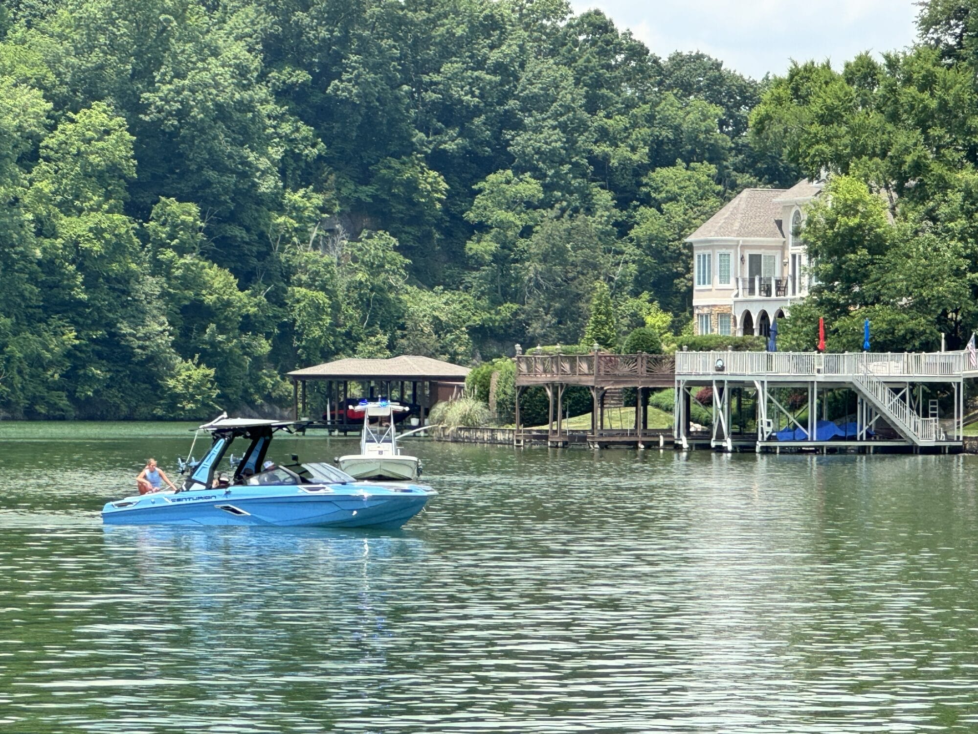 A blue motorboat with several people onboard cruises on a lake near a large lakeside house with a dock and surrounding greenery.