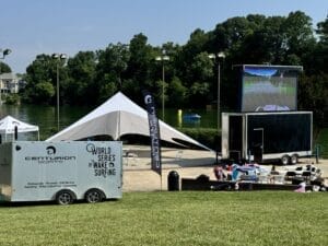 A lakeside event setup with a large tent, tables, chairs, and a trailer displaying "World Series of Wake Surfing" next to a screen playing wake surfing footage. Green lawn and trees are in the background.