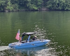 A blue motorboat with two people and an American flag cruises on a calm lake, with trees in the background.