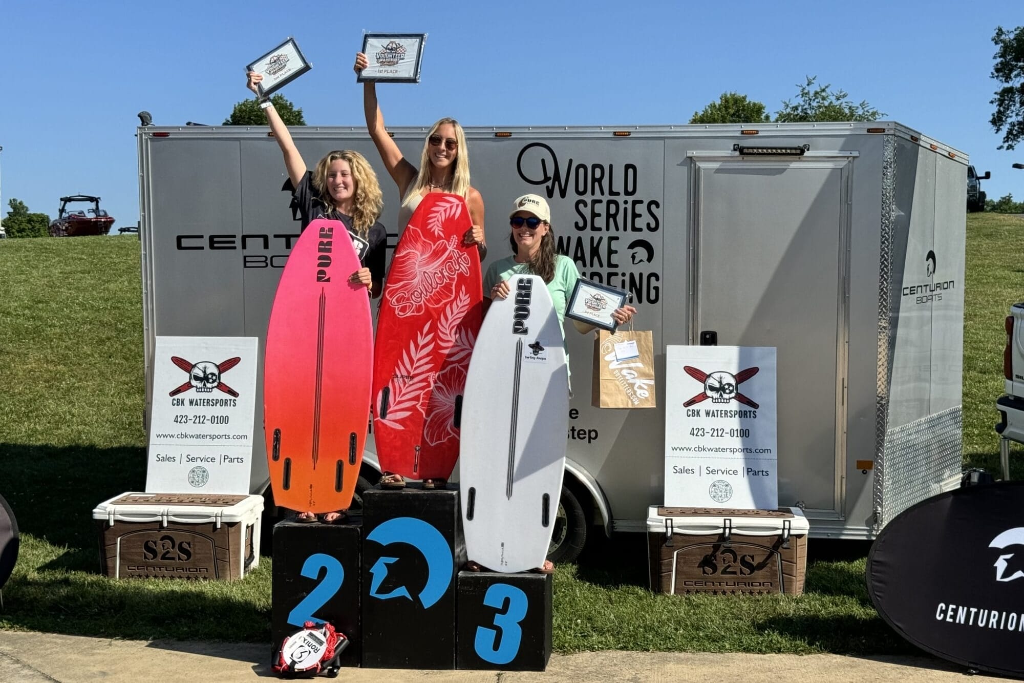 Three individuals stand on a winners podium holding certificates, with two holding surfboards. They are in front of a trailer with 