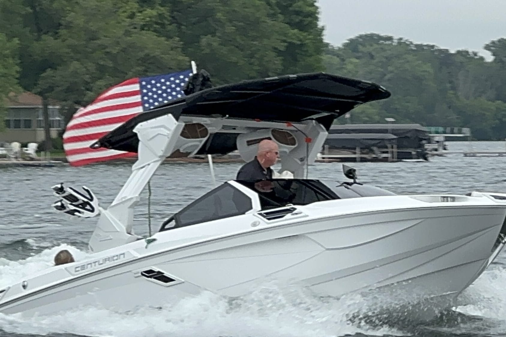 A person is driving a white Centurion boat on a lake. The boat has an American flag at the back, and there are trees and houses visible in the background.