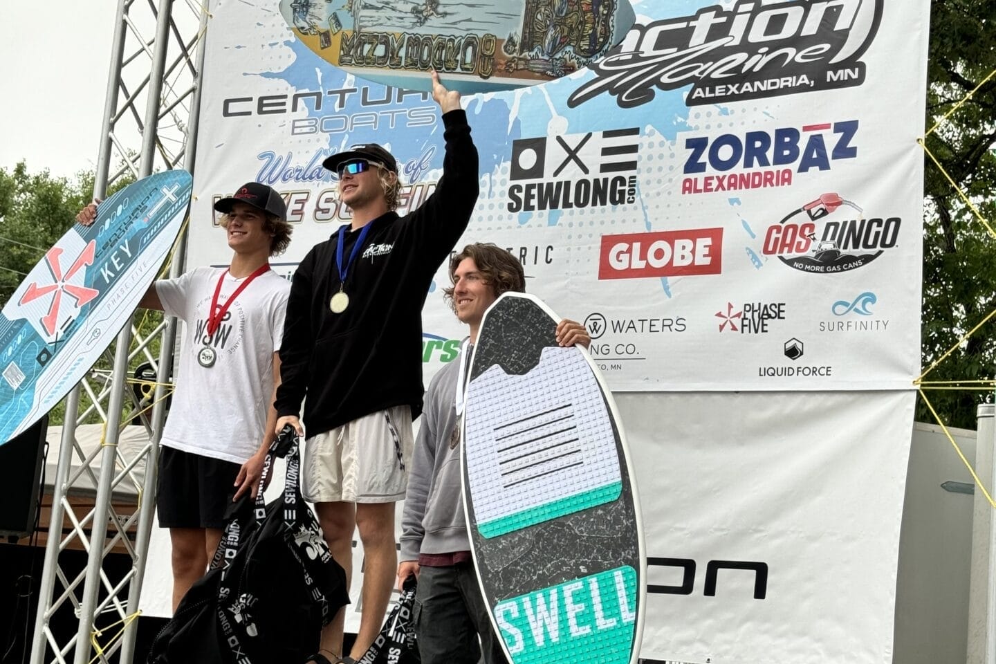 Three individuals stand on a podium holding awards at a surfing competition. The backdrop features various sponsor logos and a banner that reads 