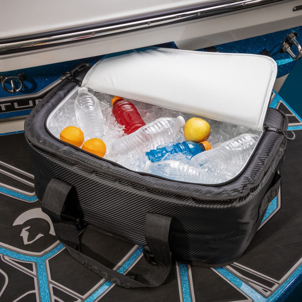 A cooler bag filled with ice, plastic bottles of water, sports drinks, and oranges, placed on a boat's deck. The lid of the cooler is partially open.