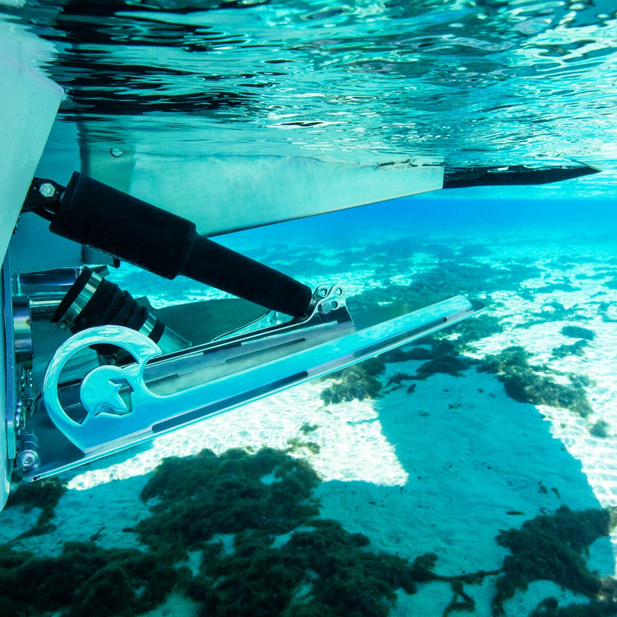 Underwater view of a boat's rudder system and propeller in clear, blue water, with the sea floor and some aquatic plants visible in the background.