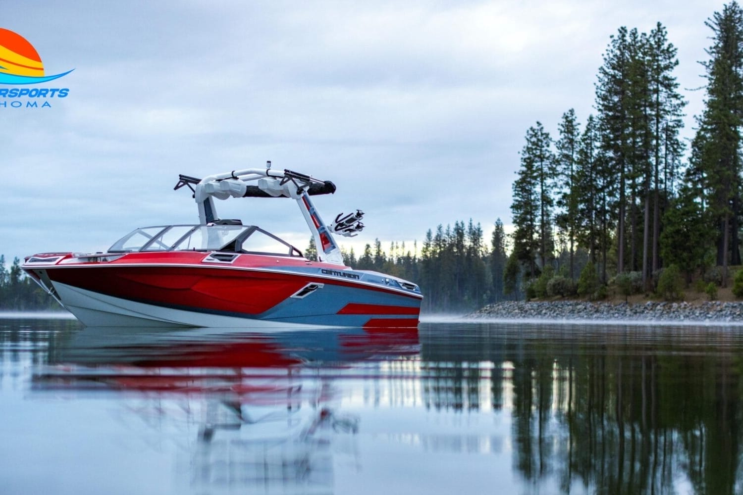 A red and white wakeboard boat is on a calm lake surrounded by trees, with a logo of 