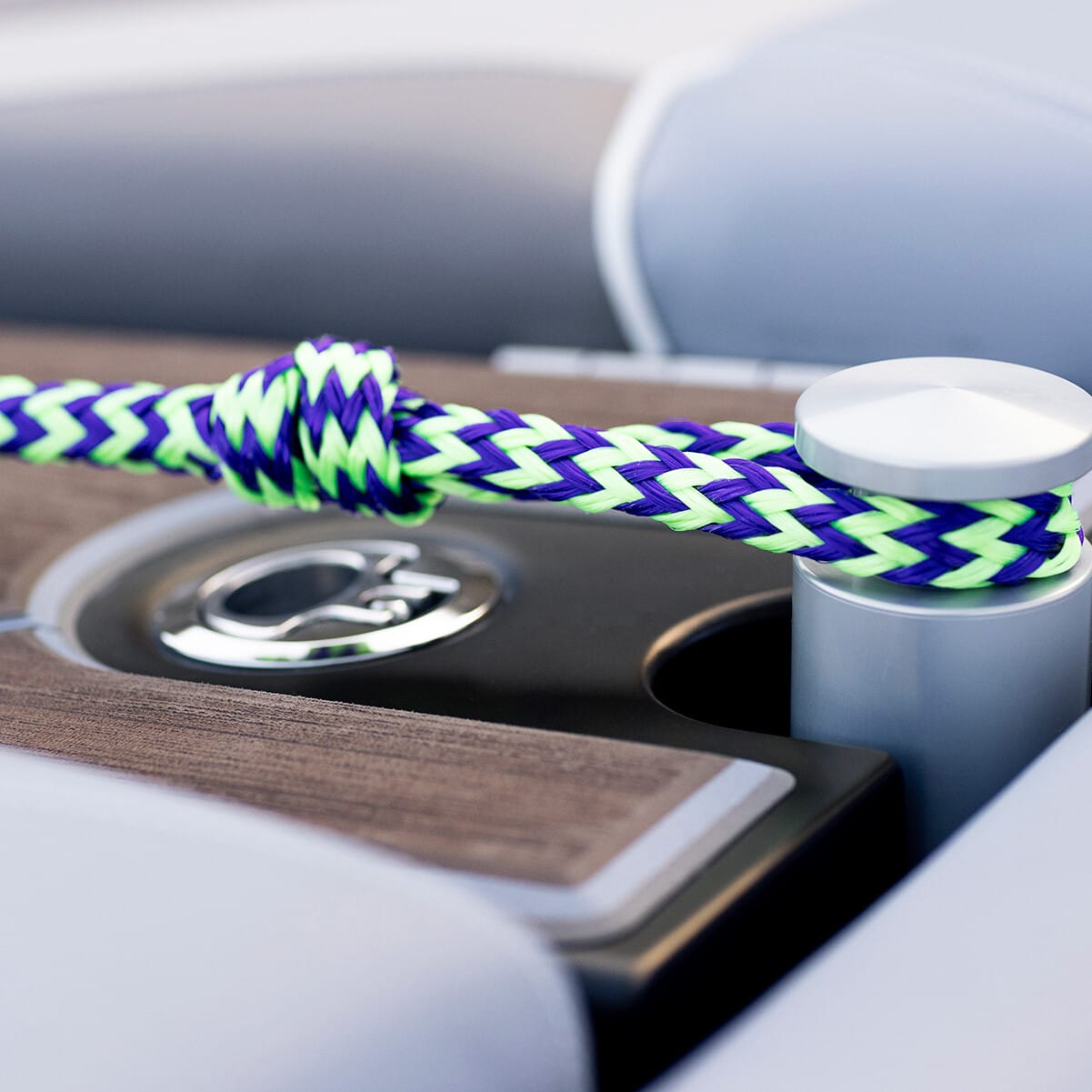 A purple and green braided rope is secured to a boat's cleat, situated between light gray cushioned seats and a wooden deck area.