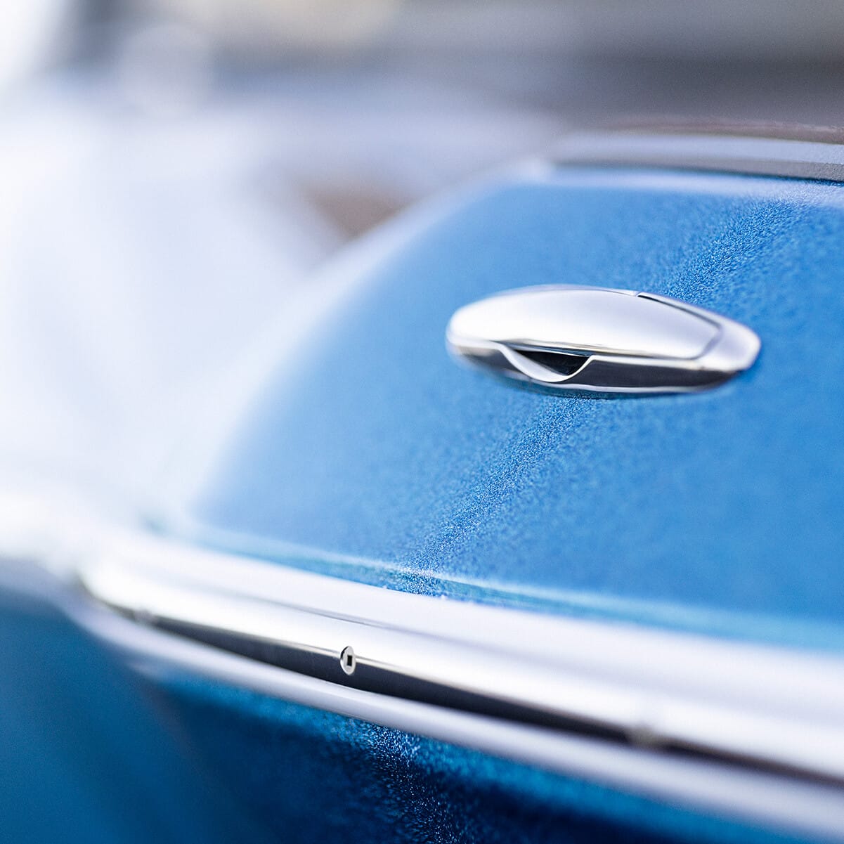 Close-up of a classic car's metallic blue door, featuring a chrome door handle and trim. The background is blurred.
